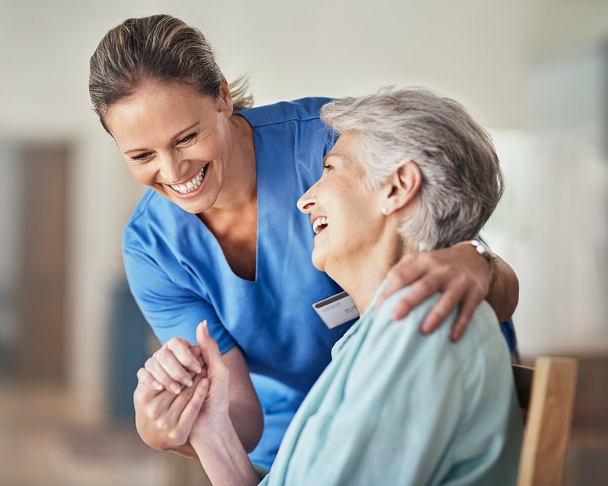 Pflegerin und Seniorin halten sich an der Hand während sie gemeinsam lachen.