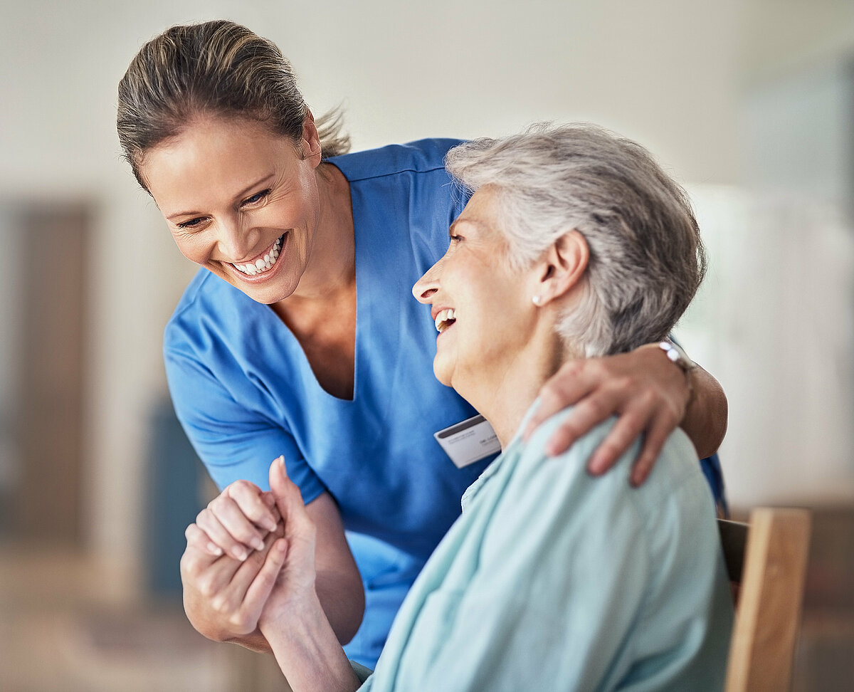 Pflegerin und Seniorin halten sich an der Hand während sie gemeinsam lachen.